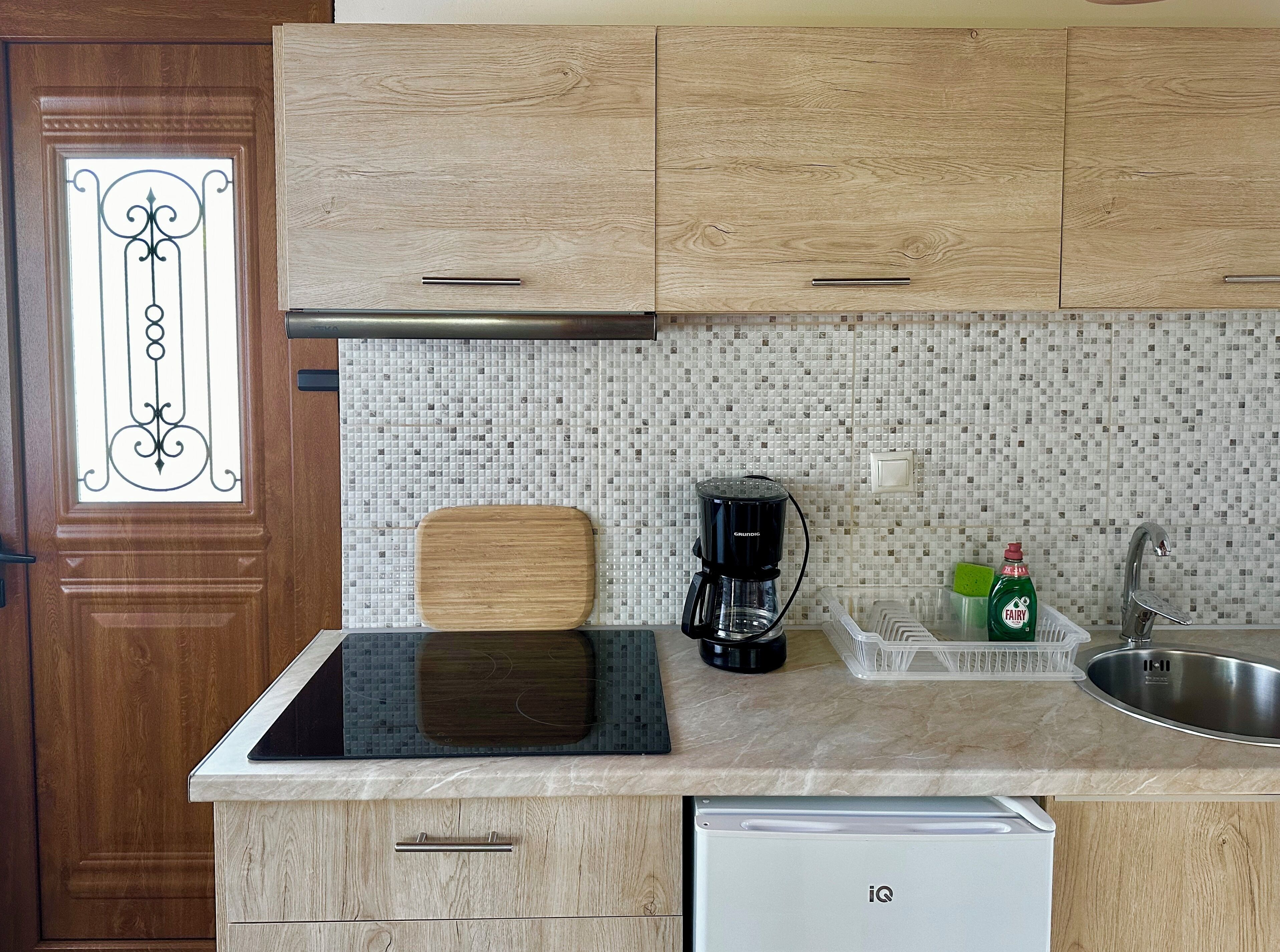 Una cocina con gabinetes de madera, salpicadero de azulejos, cafetera, tabla de cortar, escurreplatos y lavavajillas.