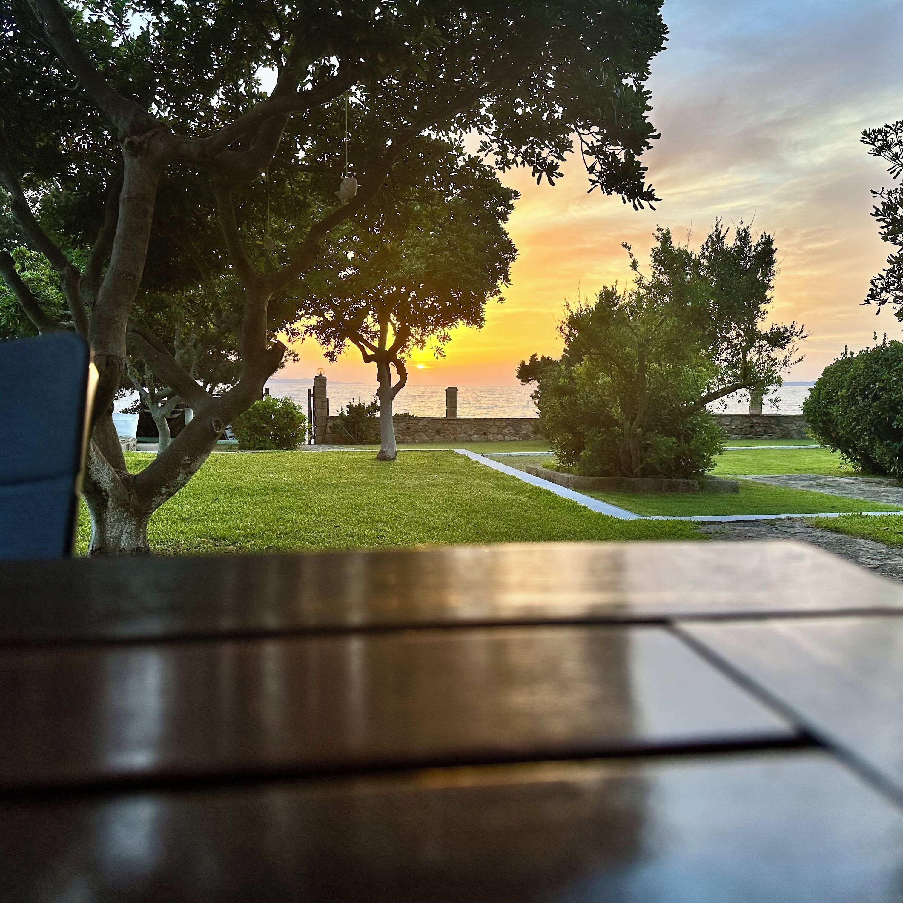 A table with a view of a sunset over a body of water, surrounded by trees and a stone wall.