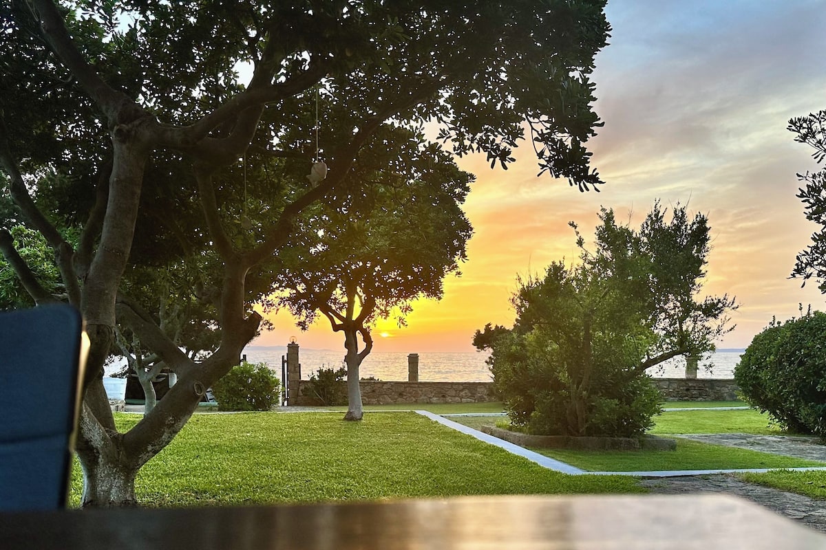 A patio table with a view of a sunset over a body of water, surrounded by trees and a stone wall.