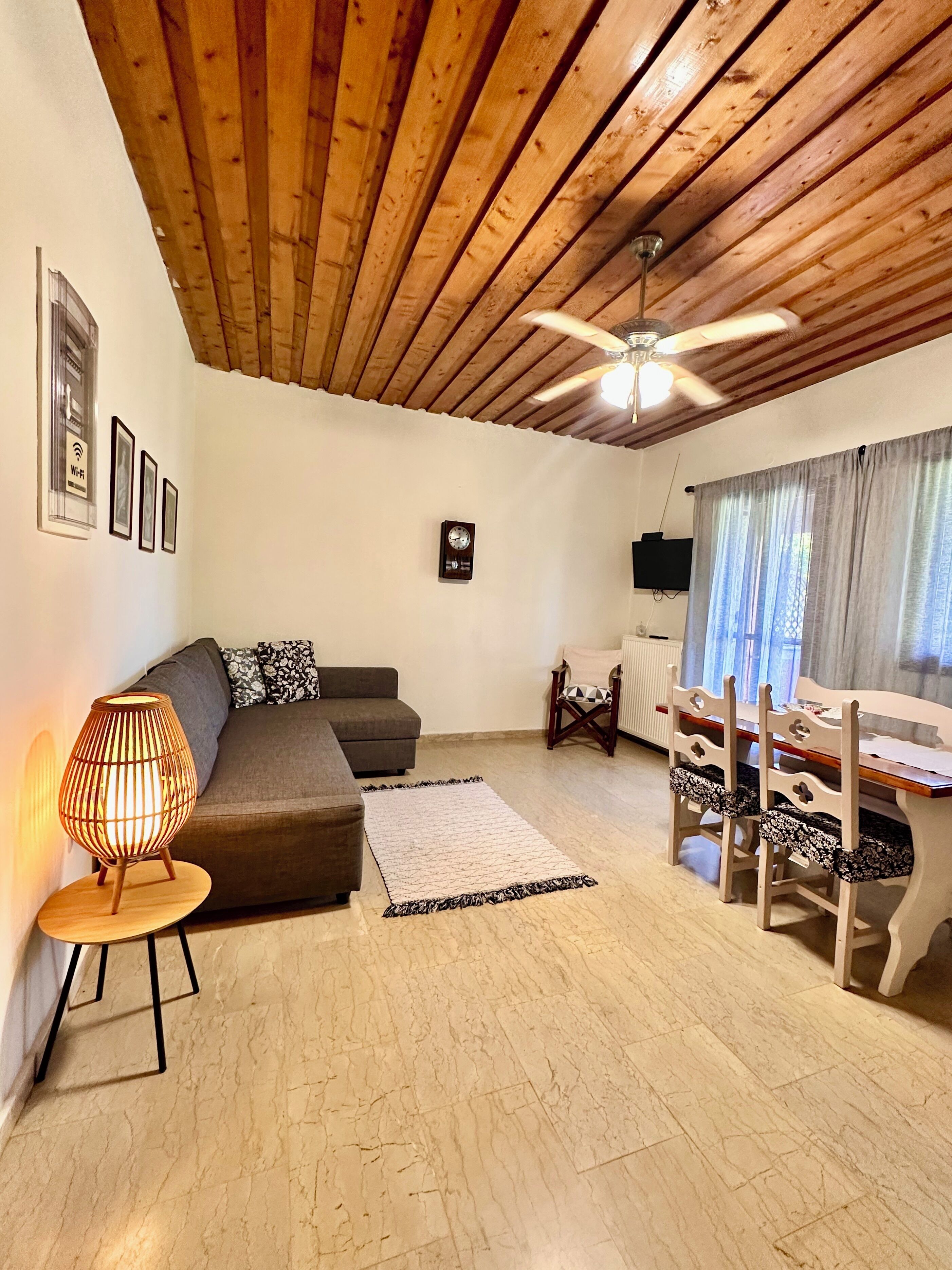 Una sala de estar acogedora con techo de madera, un sofá, una mesa con sillas, una lámpara y cuadros enmarcados en la pared.
