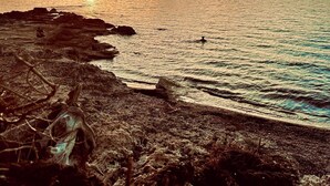 Un paisaje costero con un atardecer sobre el océano, una costa rocosa y una persona sentada sobre las rocas.