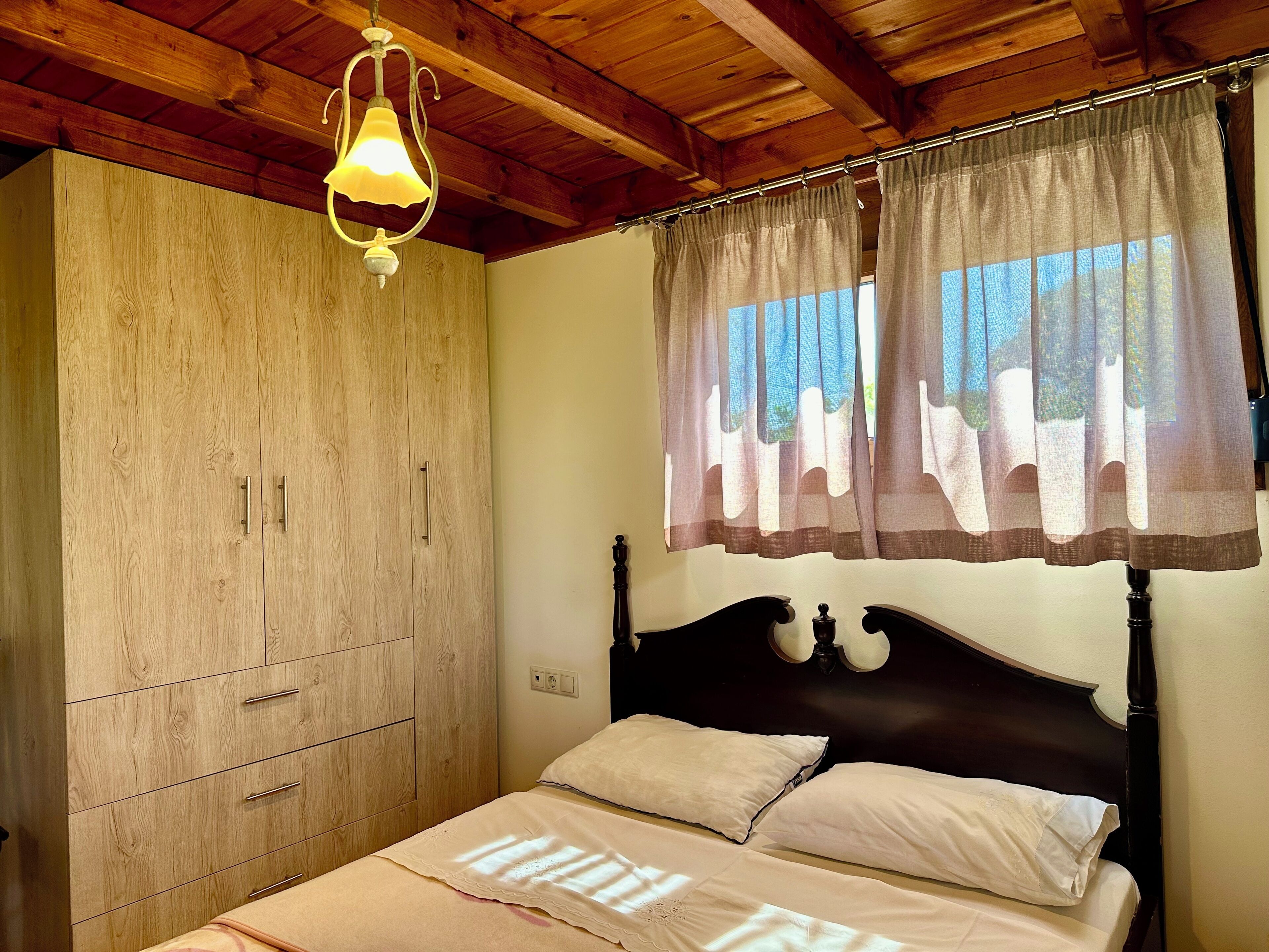 A bedroom with a wooden wardrobe, a bed with white bedding, and a window with sheer curtains.