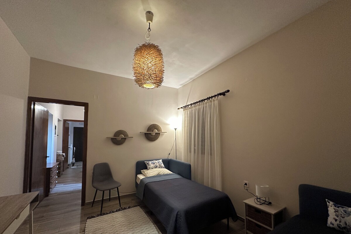 Quarto com cama, mesas de cabeceira, uma cadeira, escrivaninha, luminária pendente e janela com cortinas.