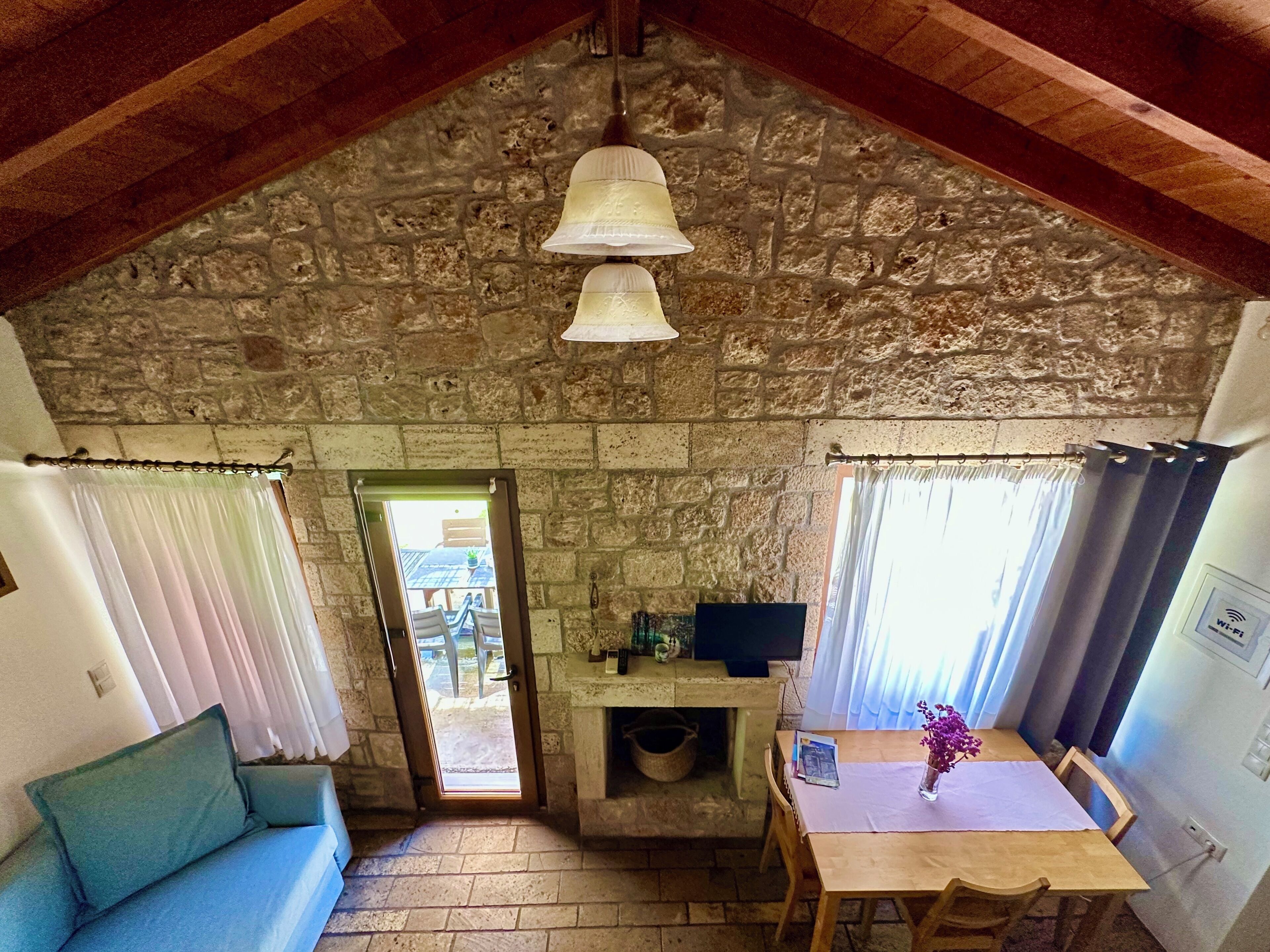 Une pièce chaleureuse avec un plafond en pierre, un plafond en bois, une table à manger avec des chaises, un canapé, un téléviseur et une fenêtre avec des rideaux.