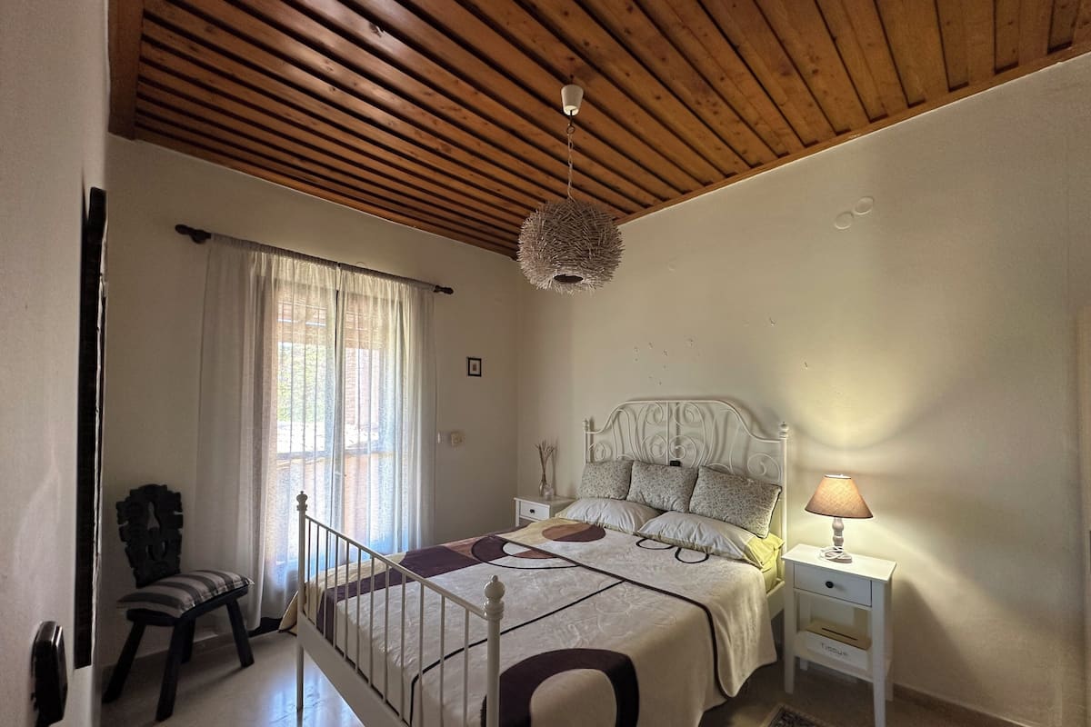 A bedroom with a wooden ceiling, a bed with a white headboard, a bedside table with a lamp, and a chair.