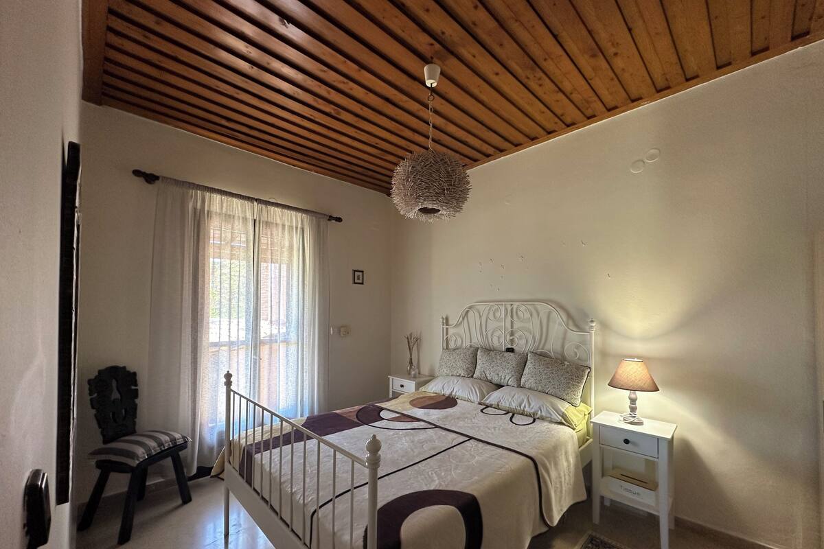 A bedroom with a wooden ceiling, a bed with a white headboard, a bedside table with a lamp, and a chair.