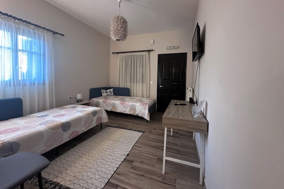 A small, well-lit room with two single beds, a desk, a chair, and a television mounted on the wall.