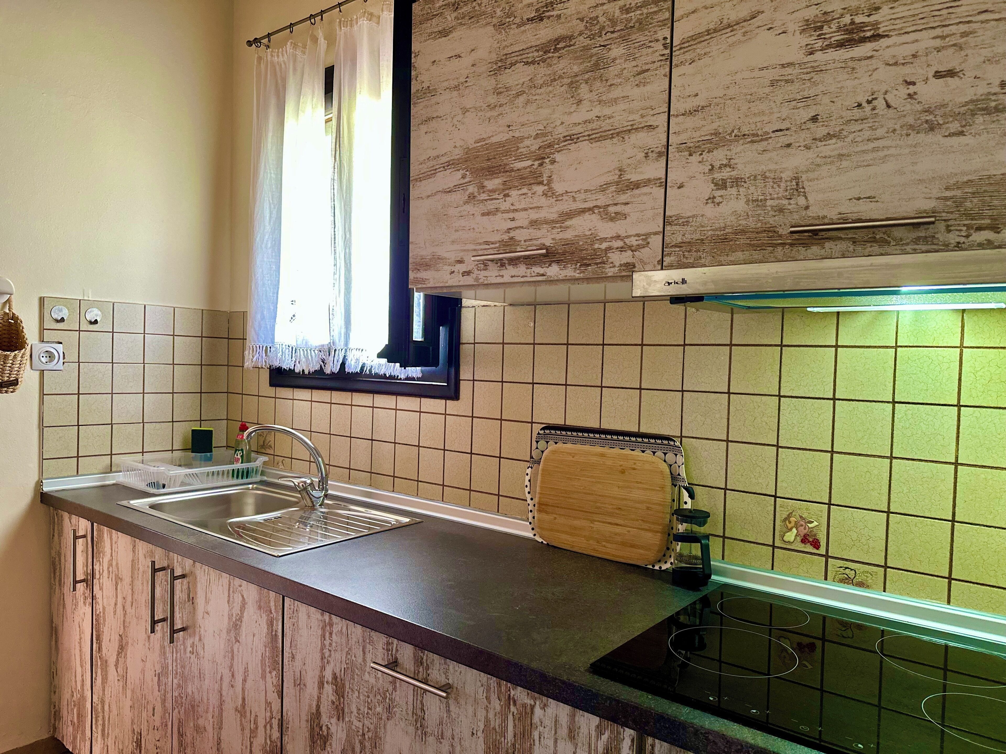 Une cuisine équipée d’un évier, de meubles en bois, d’une fenêtre avec des rideaux et d’un plan de travail recouvert de carreaux.