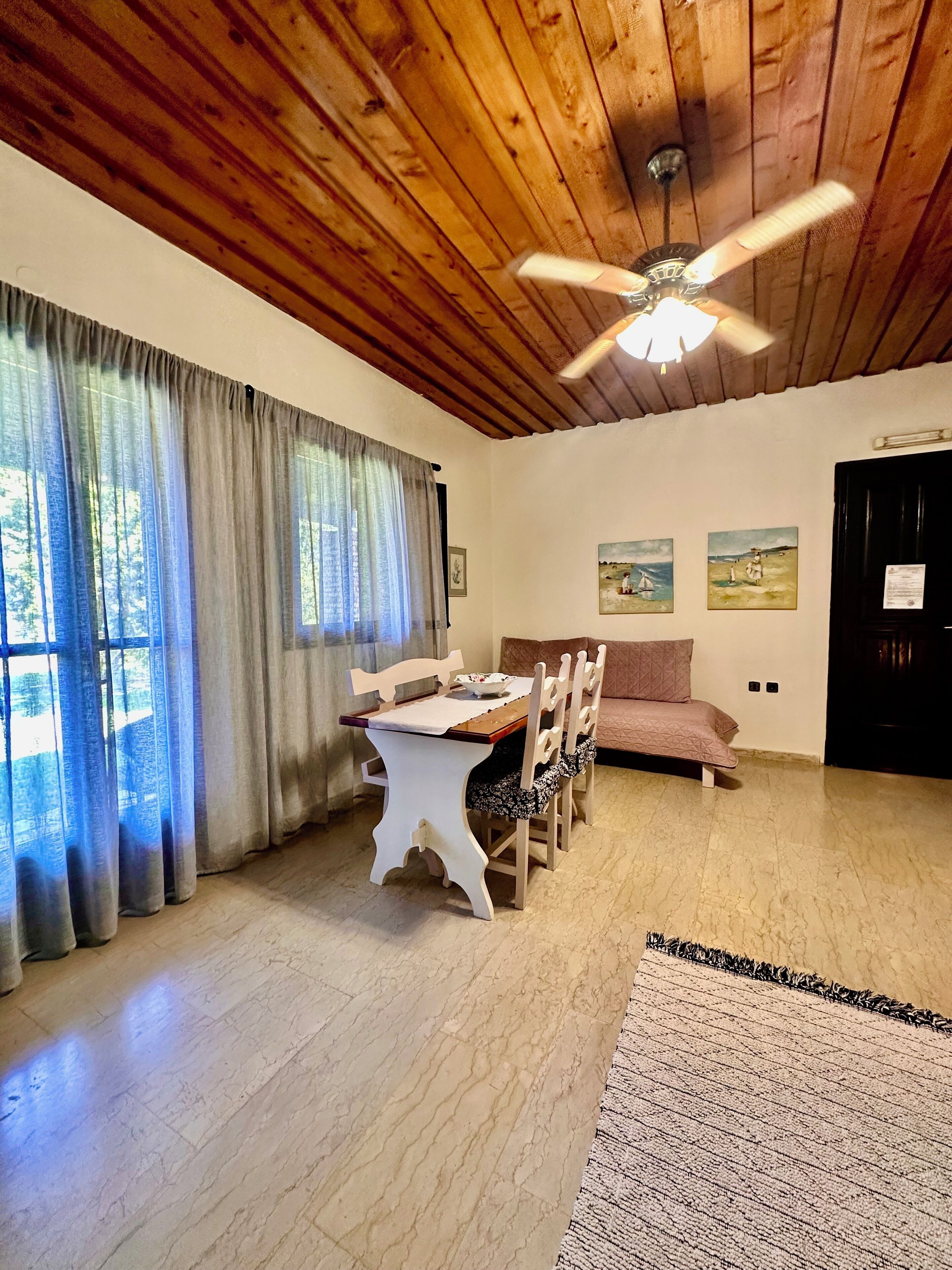 A dining area with a wooden table and chairs, a ceiling fan, and a view through large windows with curtains.