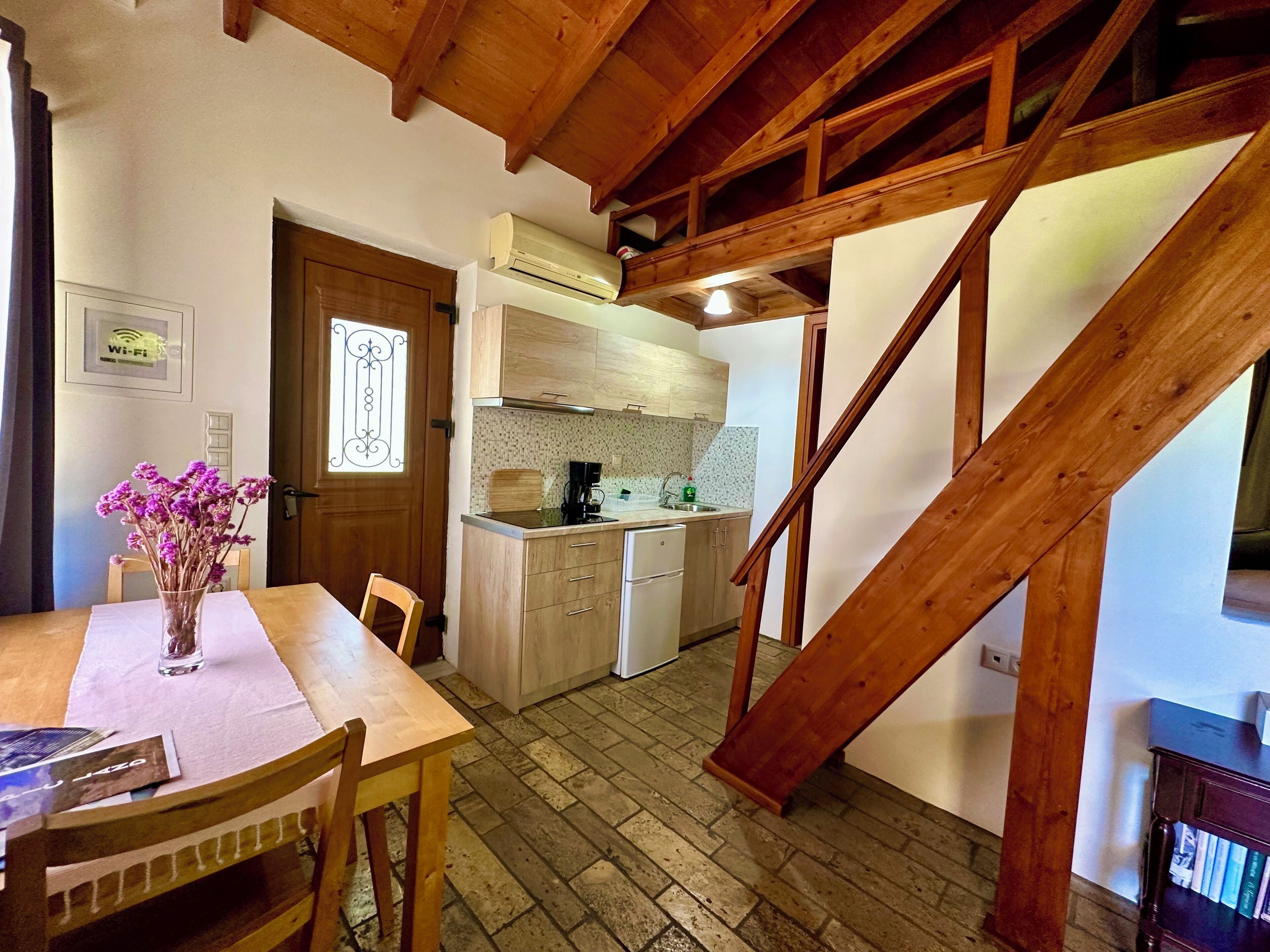 A kitchen with wooden cabinets, a coffee maker, and a sink. There is a wooden table with chairs and a vase of flowers. A staircase is visible in the background.