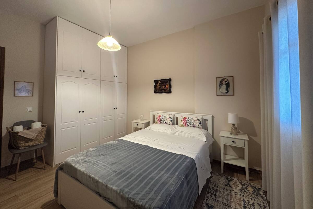 A bedroom with a bed, bedside tables, a wardrobe, a hanging light fixture, and a chair.