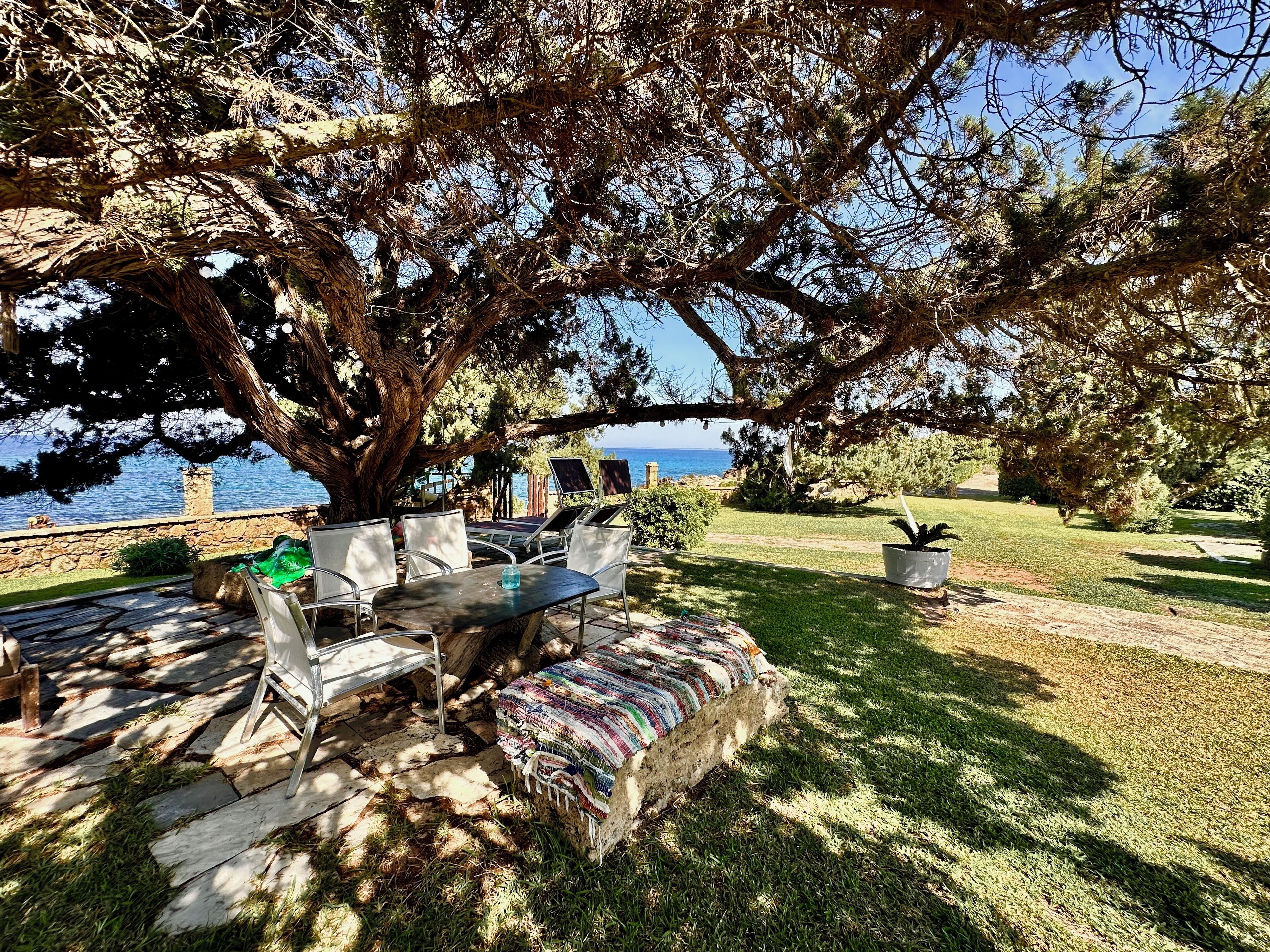 An outdoor seating area with a table and chairs, surrounded by trees and a view of the sea.