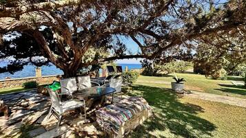 An outdoor seating area with a table and chairs, surrounded by trees and a view of the sea.