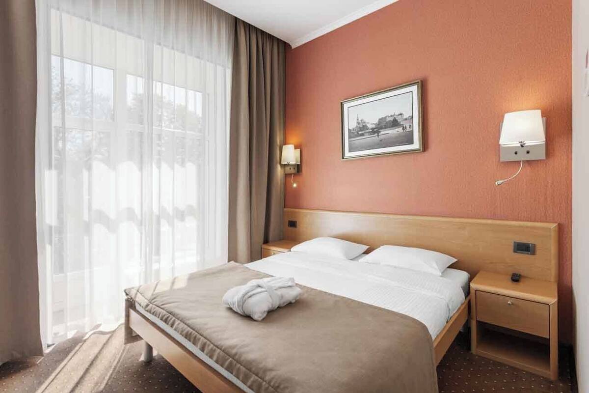 A hotel room with a bed, a nightstand, a wall-mounted lamp, a framed picture, and sheer curtains.