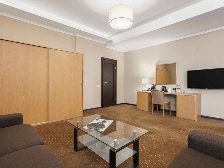 Habitación de hotel con sofá, una mesa de centro de vidrio, escritorio con silla, televisión y armario.
