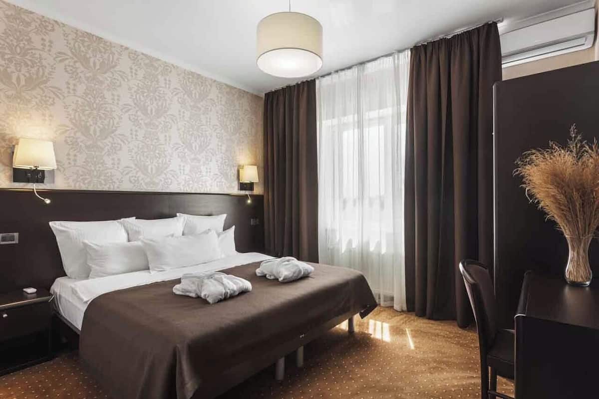 A hotel room with a large bed, two bedside lamps, a desk with a chair, and a vase with dried plants.