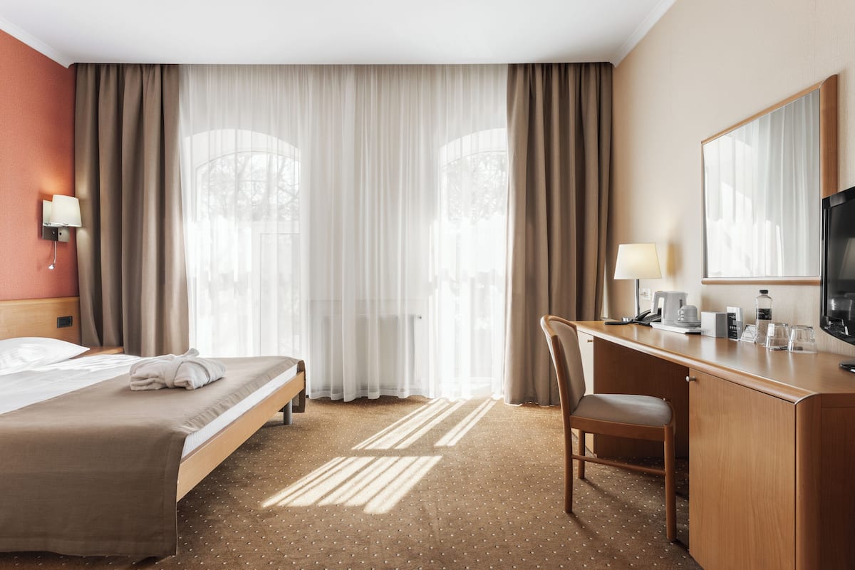 A hotel room with a bed, desk, chair, and large windows with curtains.
