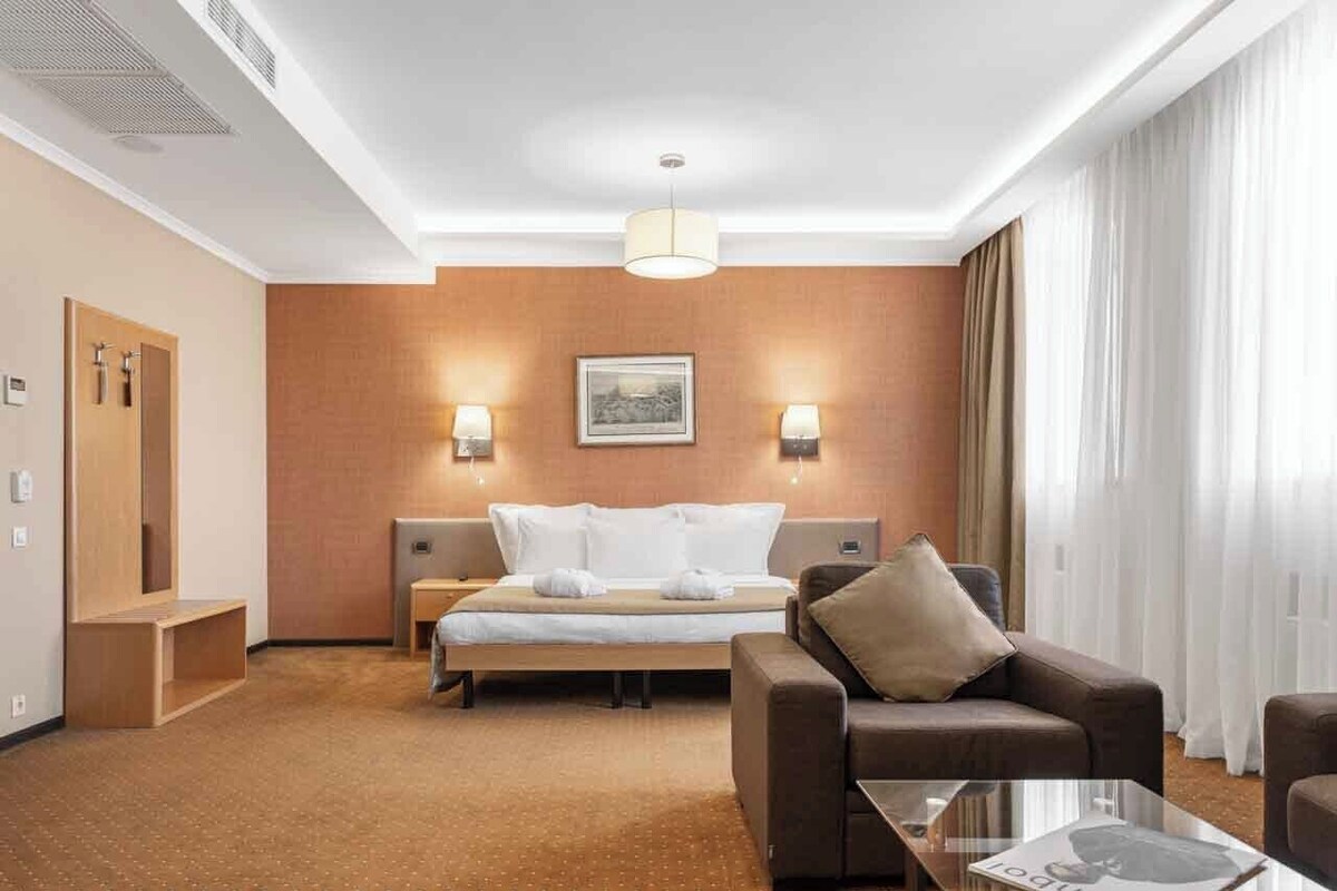 Una habitación de hotel con una cama, dos mesitas de noche, un sofá, una mesa de centro y un ventanal con cortinas.