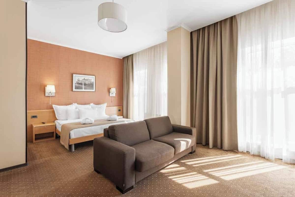 A hotel room with a bed, a sofa, a wall-mounted lamp, a picture, and a large window with curtains.