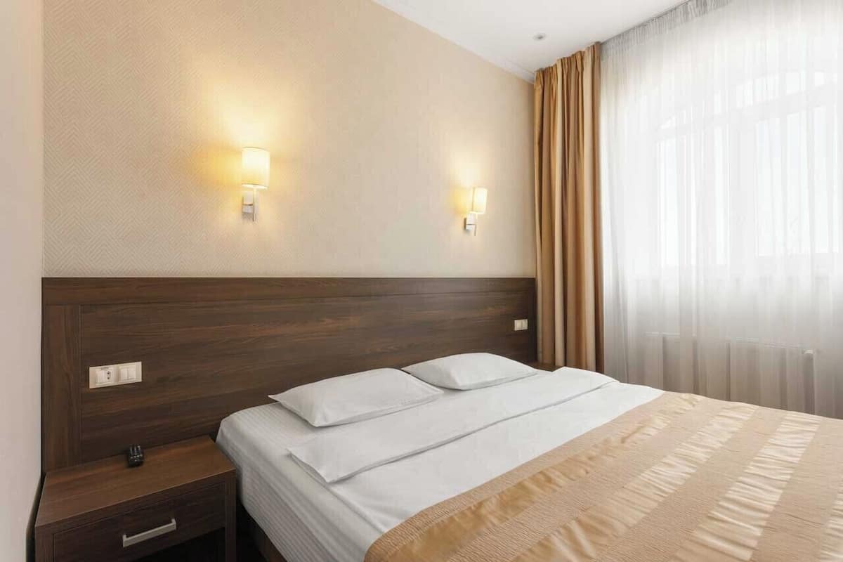 A hotel room with a wooden headboard, two bedside lamps, a bed with white linens, a nightstand with a drawer, and sheer curtains on the window.