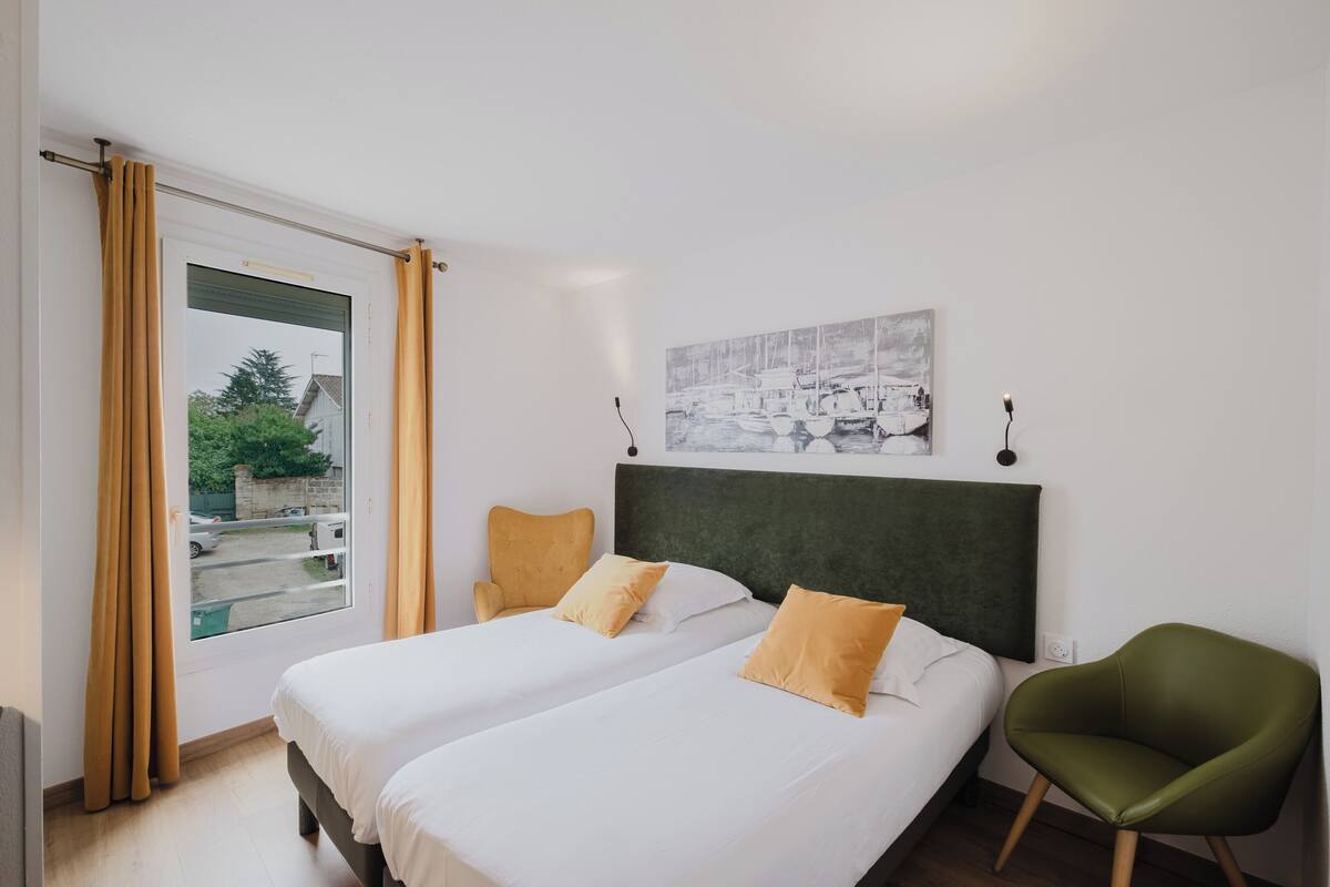 A hotel room with two beds, a green chair, a window with curtains, and a wall-mounted picture.