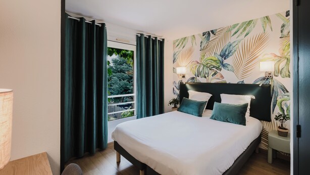 A bedroom with a large bed, dark curtains, a window with a view of greenery, and a tropical-themed wall mural. - Hôtel Amaya (Ambares-et-Lagrave)
