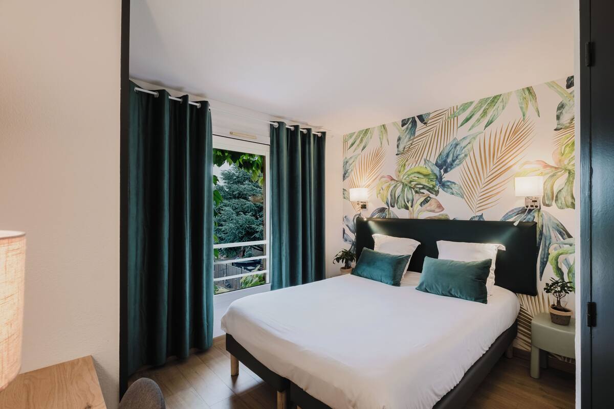 A bedroom with a large bed, dark curtains, a window with a view of greenery, and a tropical-themed wall mural.