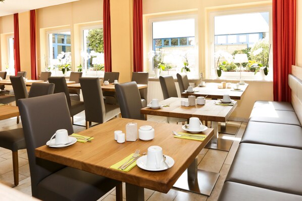 Un comedor de hotel con varias mesas y sillas, dispuesto en un espacio amplio con ventanas grandes que permiten la entrada de luz natural.