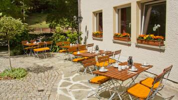 Eine Terrasse im Freien mit Tischen und Stühlen, einem Steinboden und Blumenkästen an den Fensterbänken des Gebäudes.