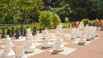 Un grand échiquier extérieur, avec des pièces blanches et noires disposées sur une surface pavée, entouré de verdure et d’arbres.