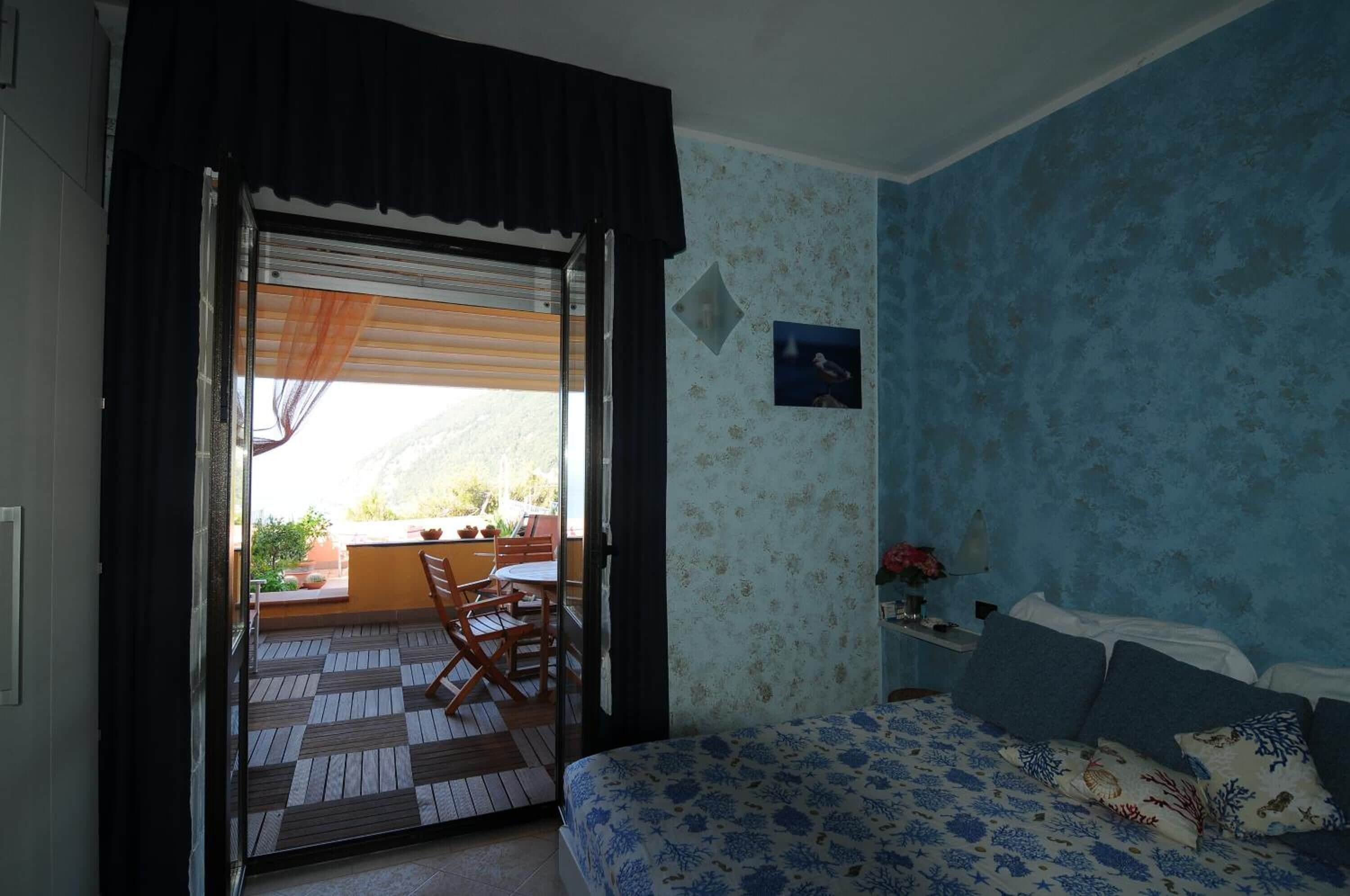 Un dormitorio con cama, mesita de noche, ventana con vistas al exterior y puerta que da a un espacio exterior con mesa y sillas.