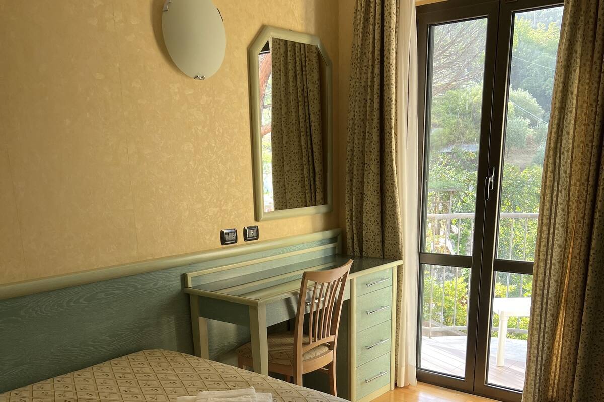 A hotel room with a bed, a desk, a chair, a mirror, and a view of greenery through the window.
