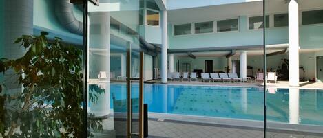 A modern indoor swimming pool with clear glass walls, lounge chairs, and a central column structure.