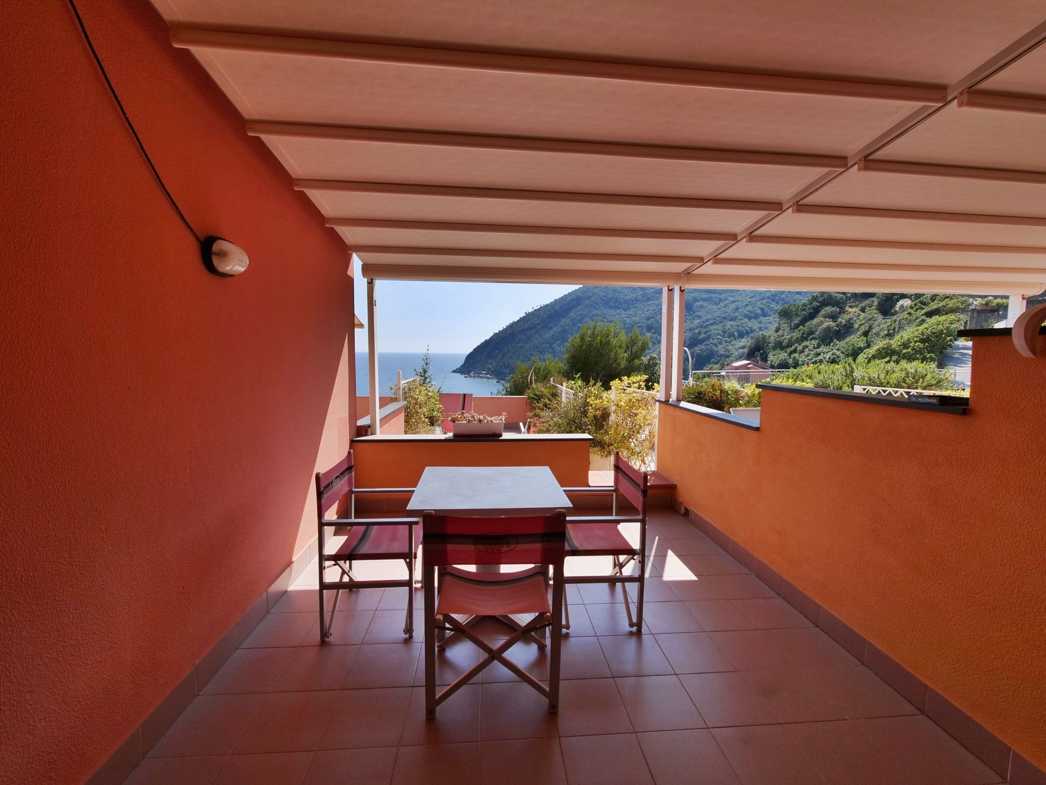 Un balcón con mesa y sillas, vista al mar y a las montañas, y una pared de color rojo.