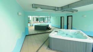 Un moderno espacio de spa con una bañera de hidromasaje grande, una zona de relajación con sillones, y una piscina visible al fondo.