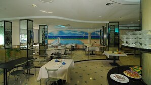 Un comedor amplio con varias mesas y sillas, un mural de una escena de playa en la pared, y una barra de buffet con diversas opciones de comida.