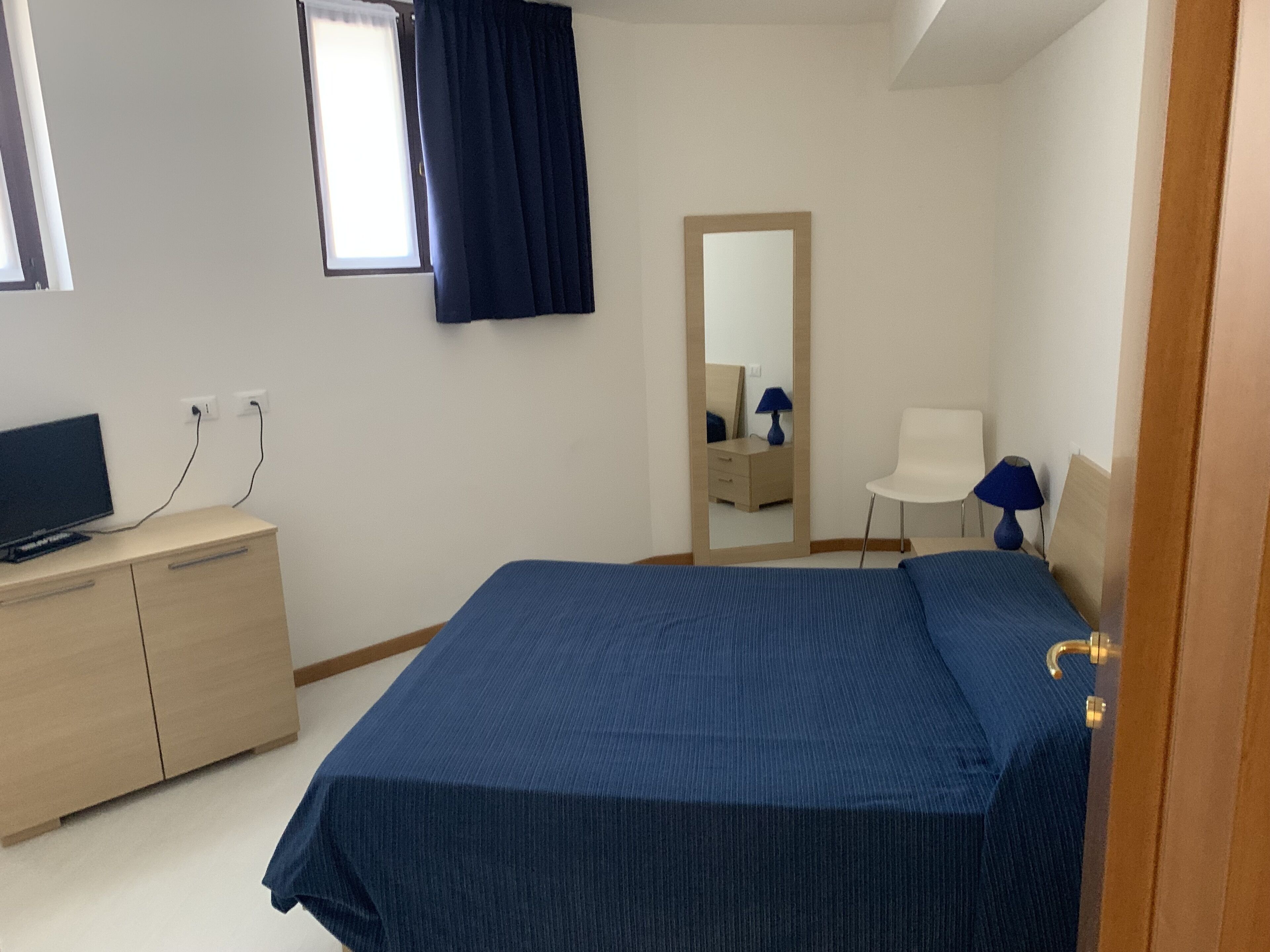 A bedroom with a bed, a desk, a chair, a mirror, and a television.