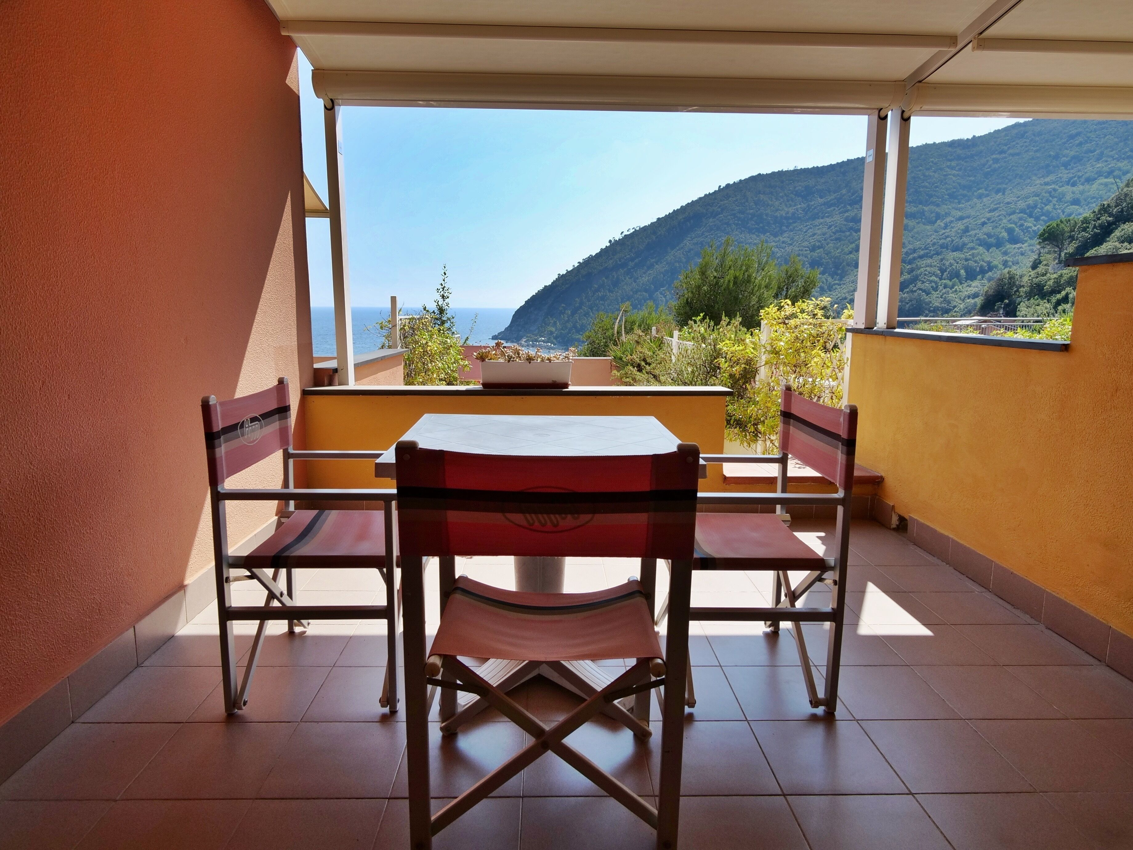 A balcony with a table and chairs, a view of the sea and mountains, and a yellow wall.