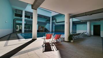 A spacious indoor pool area with a clear view of the pool, lounge chairs, and a reception desk in the background.