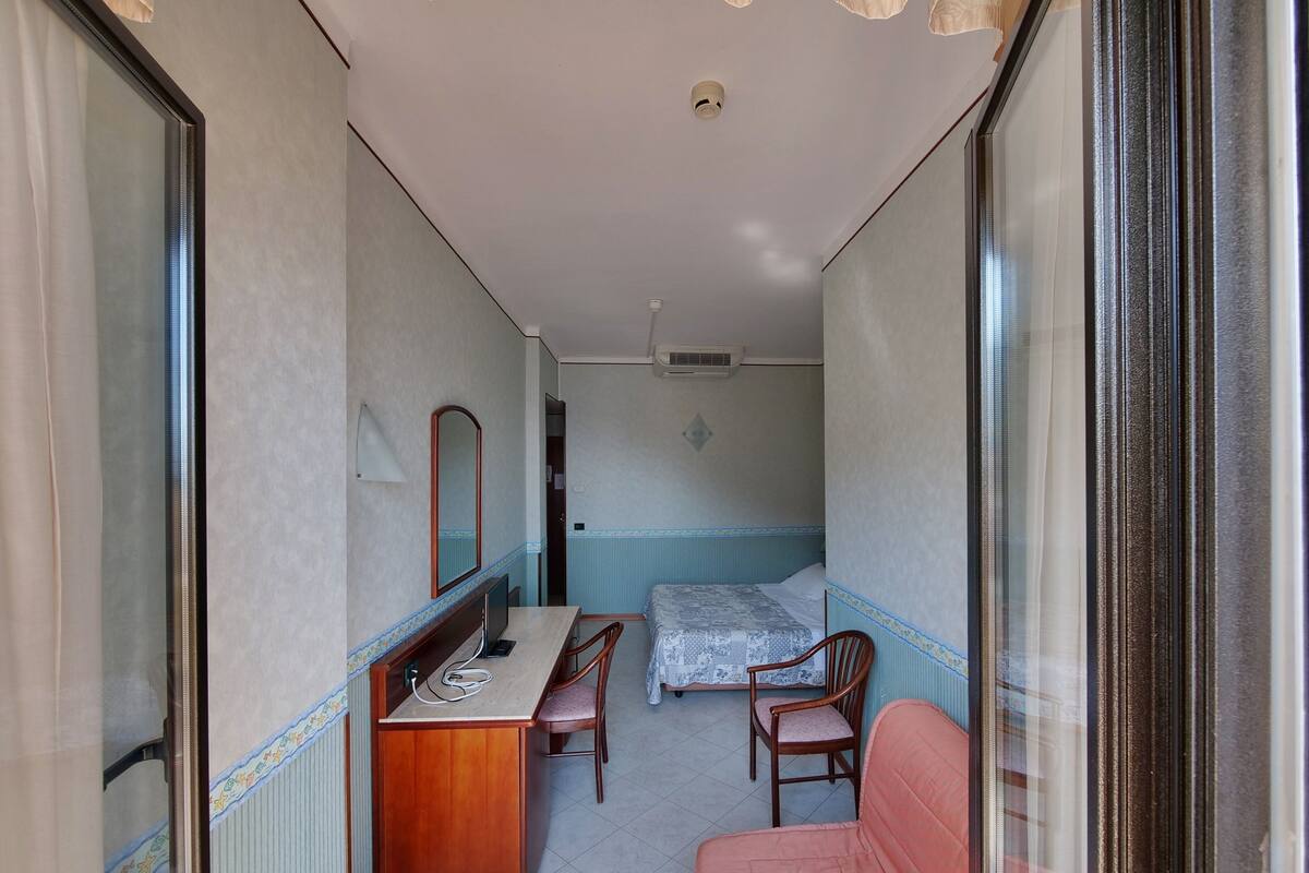 A compact hotel room with a single bed, a desk with a chair, a sofa, and a mirror.
