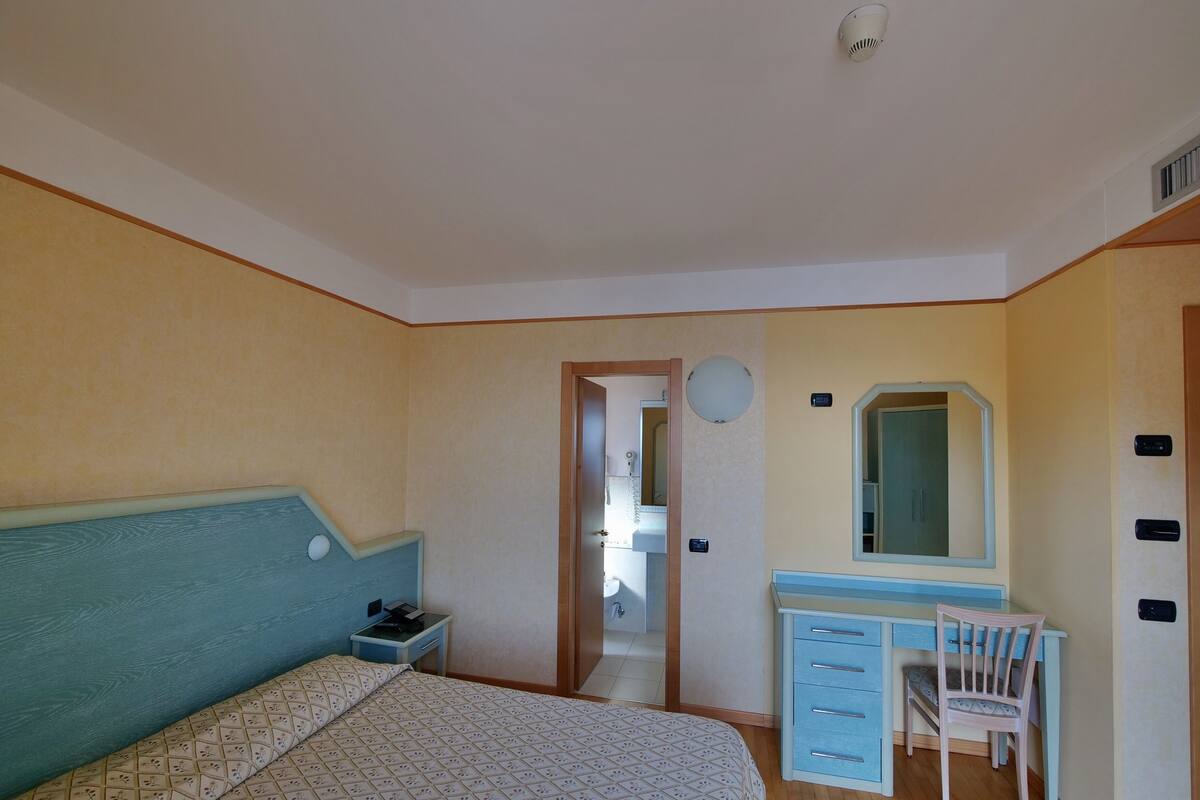 A bedroom with a bed, a desk with a chair, a mirror, and a door leading to a bathroom.