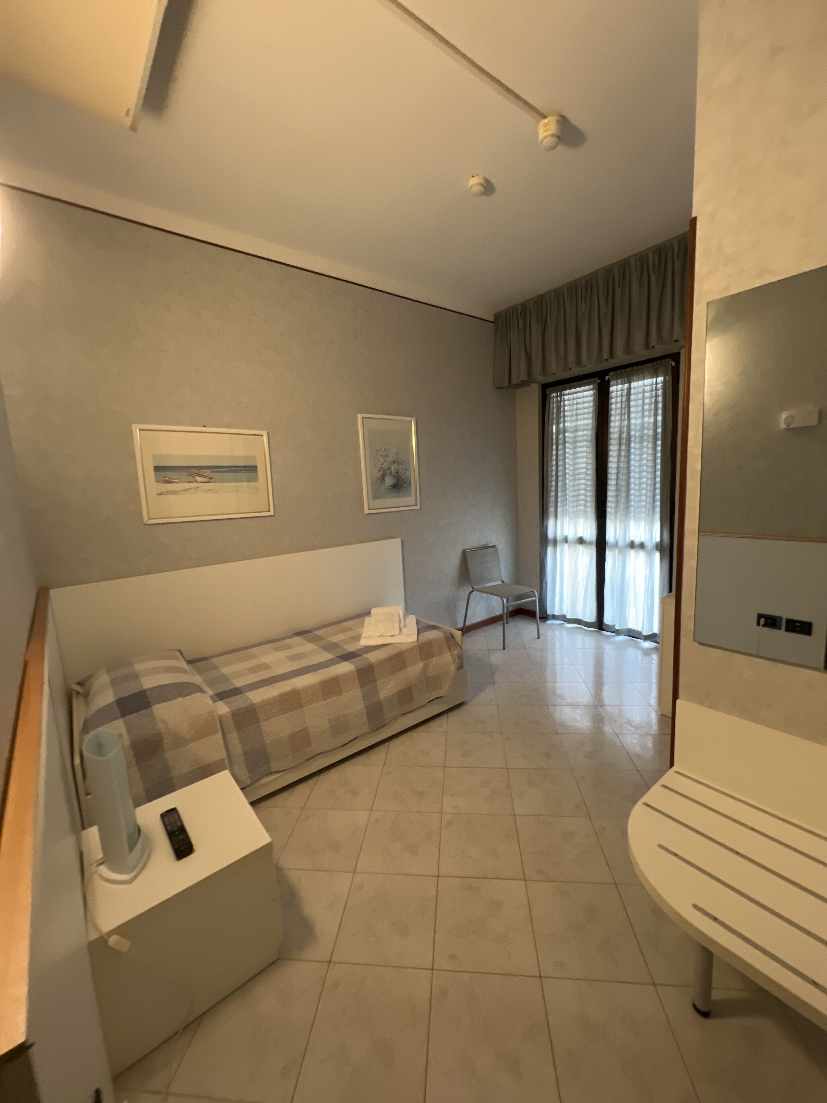 Un dormitorio con cama, una silla, un escritorio con lámpara y un ventanal con cortinas.