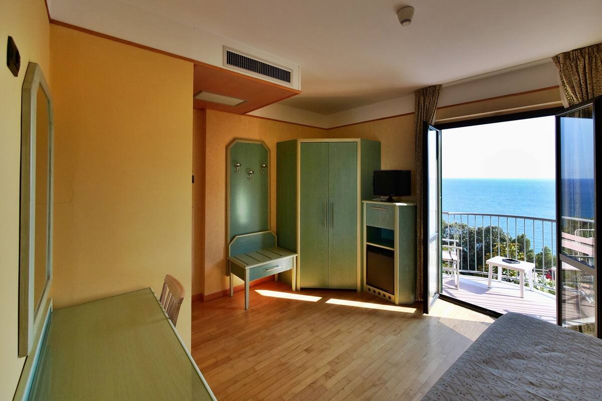 A room with a wooden floor, a green wardrobe, a small table with a TV, and a balcony with a view of the sea.