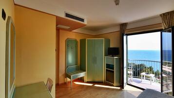 Quarto com piso de madeira, guarda-roupa verde, uma mesinha com uma TV e varanda com vista para o mar.