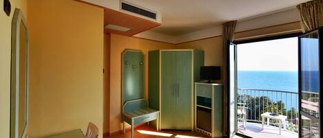 A room with a wooden floor, a green wardrobe, a small table with a TV, and a balcony with a view of the sea.