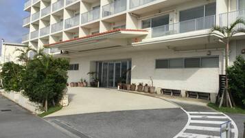 Un hotel de varios pisos con fachada plana, balcones y un área de entrada cubierta con plantas en macetas.