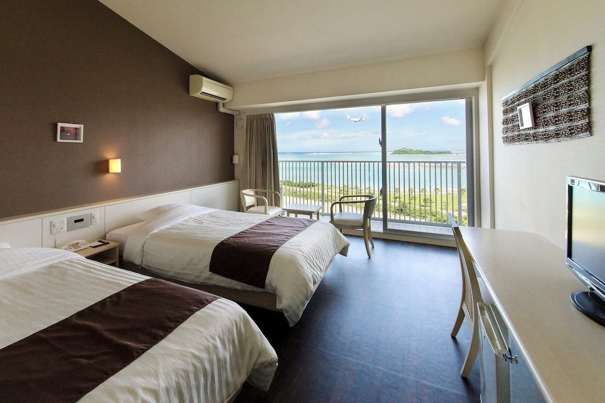 A hotel room with two beds, a desk, a TV, and a balcony with a view of the sea and a plane.