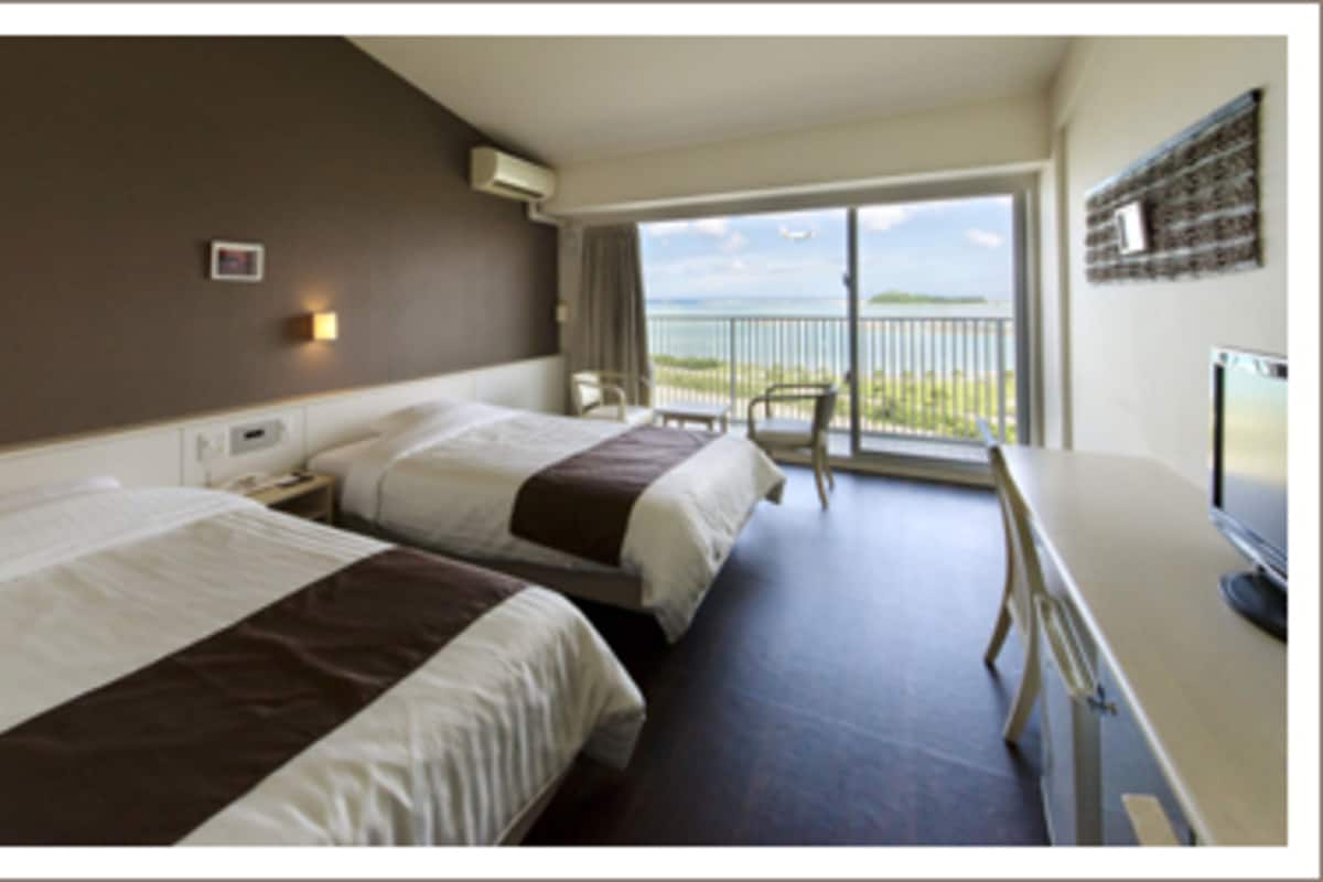 A hotel room with two beds, a desk, a chair, a TV, and a balcony with a view of the sea.