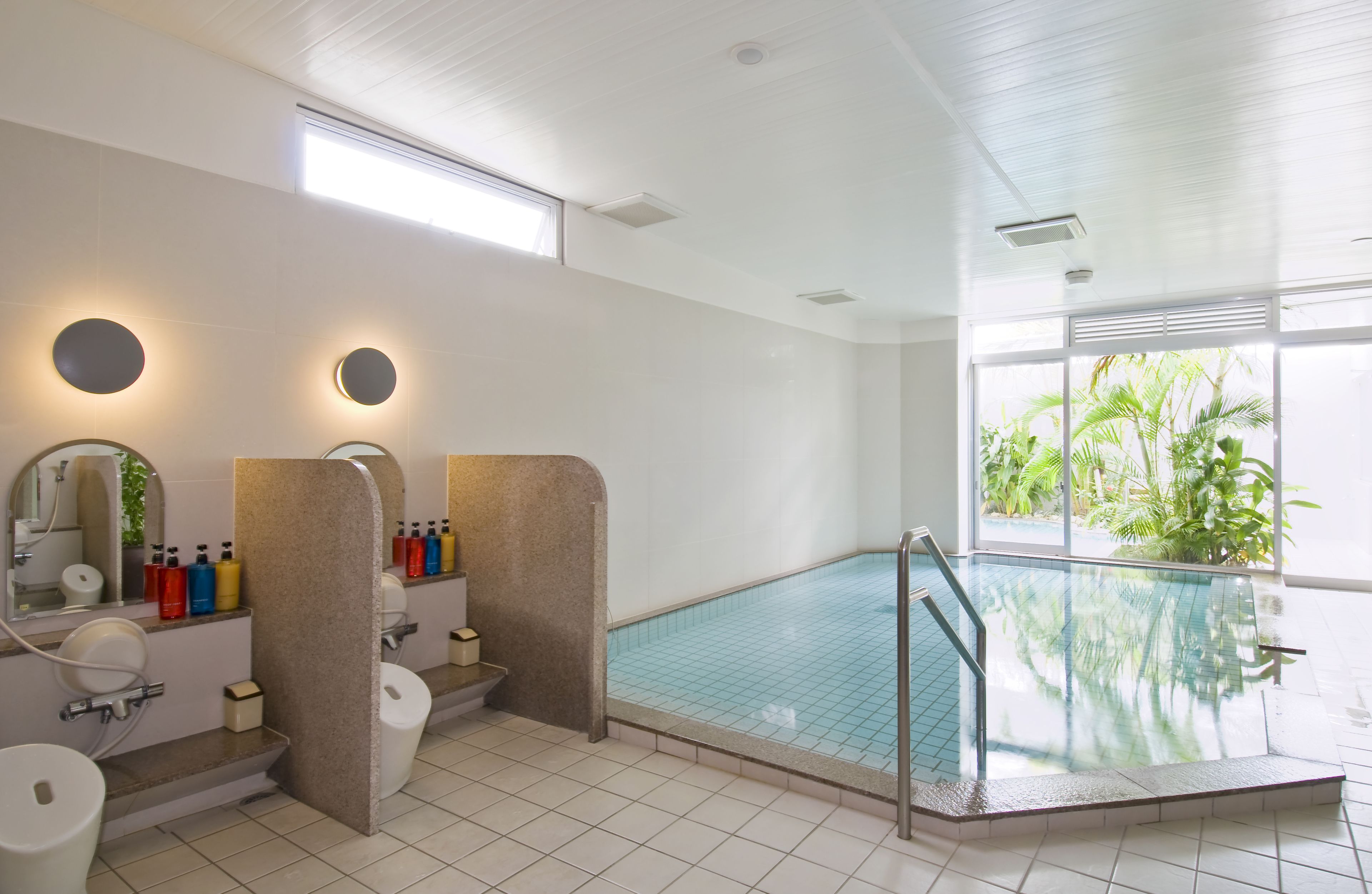 Una piscina interior moderna con pared de vidrio, un espacio iluminado por el sol con un gran ventanal, y un baño con espejo, lavamanos y artículos de tocador.