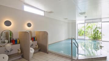 Una piscina interior moderna con pared de vidrio, un espacio iluminado por el sol con un gran ventanal, y un baño con espejo, lavamanos y artículos de tocador.